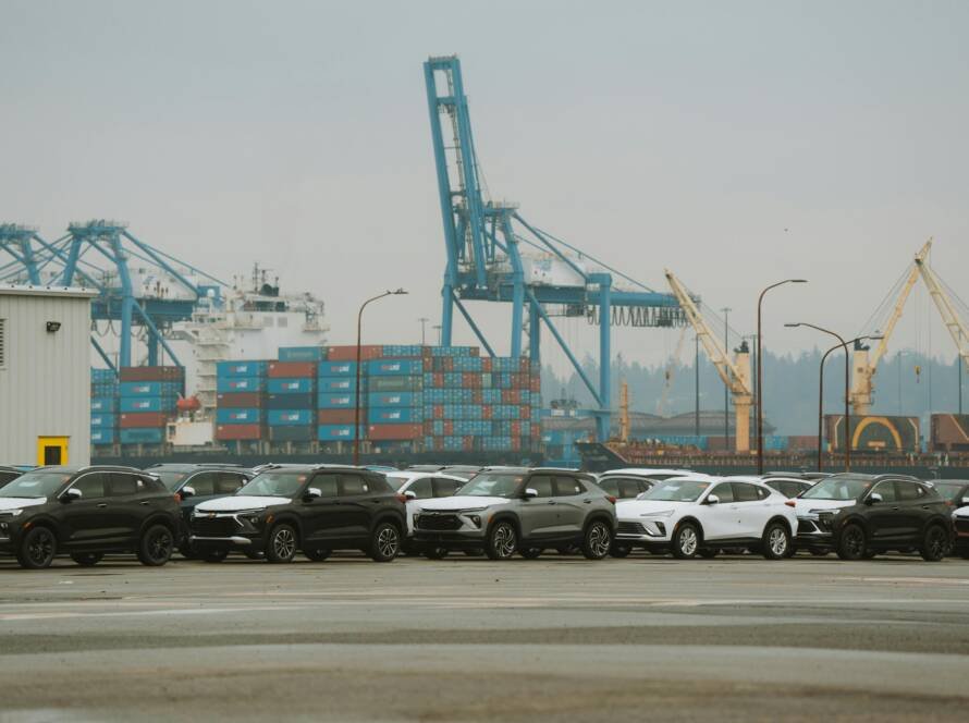A lineup of parked cars at a bustling industrial shipping port with cranes and containers.